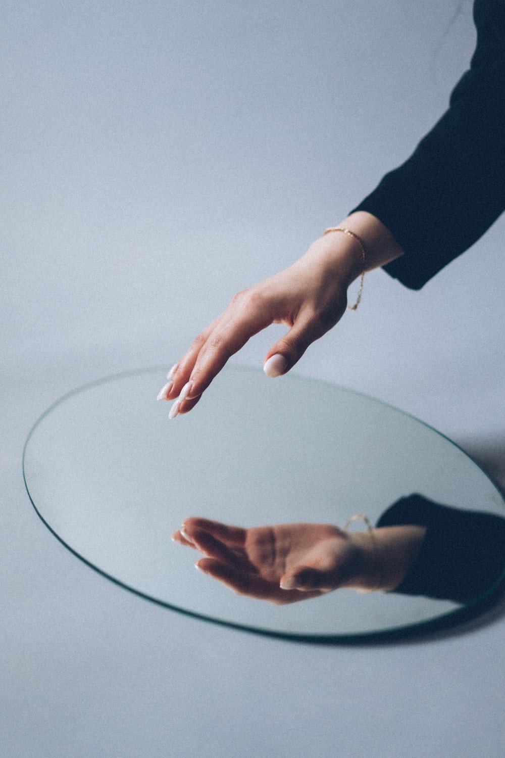 Hand with dark sleeve and gold bracelet reaching to touch a mirror with reflection of hand. 